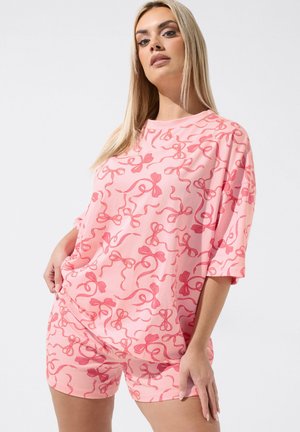 Kvinde iført pink matchende kortærmet top og shorts med røde sløjfebåndsmønstre, stående mod en hvid baggrund.