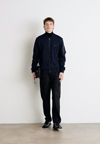 Jūras spēku zip-up jaka ar augstu apkakli, apvienota ar melniem kravas biksēm un melniem apaviem. Modelis tur nelielu somu. Minimālistisks dizains, gluda tekstūra.