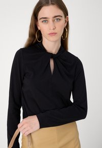 Svart langermet topp med knyttedesign ved halsen, med en nøkkelhullsutsparing. Glatt stoff med en tettsittende silhuett.