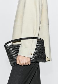 Hand hält eine schwarze gewebte Lederhandtasche mit kurzem Henkel, trägt ein cremefarbenes Langarmhemd und dunkle Hosen vor einem einfarbigen Hintergrund.