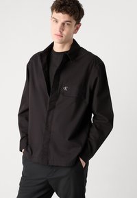 Svart skjorte med knapper foran og brystlomme, med et brodert logo. Laget av glatt stoff, har lange ermer og en klassisk kragedesign.
