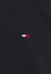 Marine tekstureret stof med et lille broderet logo med rød, hvid og blå horisontale striber på venstre side.