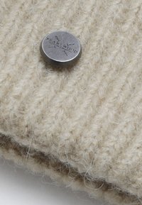 Tessuto a maglia beige con un motivo testurizzato e un bottone rotondo in metallo argentato inciso con "PEARLWOOD". Aspetto morbido e fluffy.