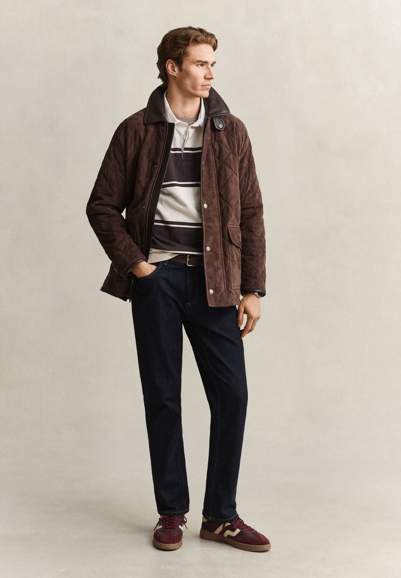 Man wearing brown quilted jacket, striped polo shirt, dark jeans, and burgundy sneakers, standing with one hand in pocket, looking right.