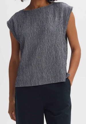Femme portant un haut sans manches gris texturé associé à un pantalon foncé, debout avec une main dans la poche devant un fond uni.