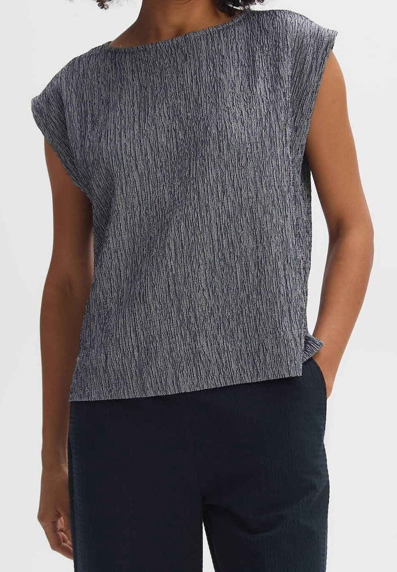 Femme portant un haut sans manches gris texturé associé à un pantalon foncé, debout avec une main dans la poche devant un fond uni.