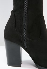 Bottine en daim noir avec talon épais de hauteur moyenne et fermeture éclair visible sur le côté.