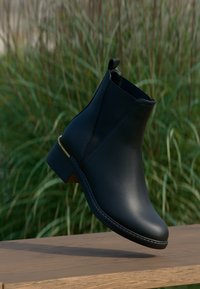 Fekete bőr bokacsizma, amelynek formája fokozatosan keskenyedik, arany díszítéssel a sarkán és sima felülettel, természetes háttér előtt elhelyezve.