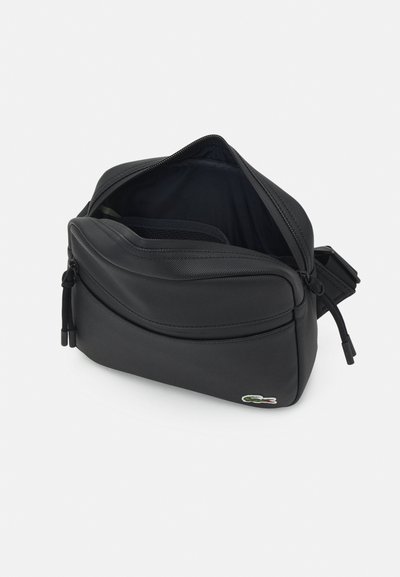 Sac en bandoulière noir en toile avec une surface texturée, fermeture éclair double, compartiments intérieurs et un petit patch logo sur le devant.