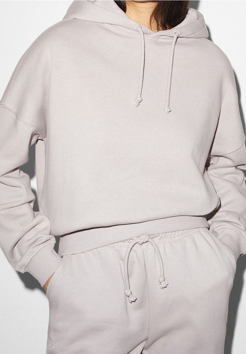 Femme portant un sweat à capuche beige clair et un pantalon de survêtement assorti, mains dans les poches, vue du cou aux hanches.