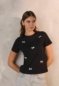 Czarny bawełniany t-shirt z białymi haftowanymi kokardkami rozsianymi po przodzie. Krótkie rękawy i okrągły dekolt. Luźny fason.