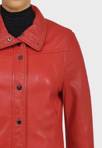 Červená kožená bunda s límečkem, plným zapínáním na knoflíky a hladkou texturou. Viditelné švy a čern é knoflíky zdobí design.