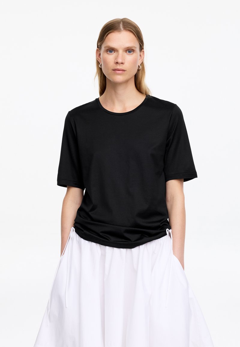 Femme aux longs cheveux blonds porte un t-shirt noir à manches courtes et une jupe blanche, debout les mains dans les poches devant un fond uni.