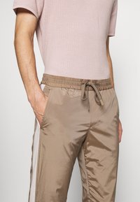 Pantalones deportivos de chándal color caqui, muy ligeros, con cintura elástica y cordón ajustable, con franjas laterales blancas en contraste y bolsillos laterales inclinados.