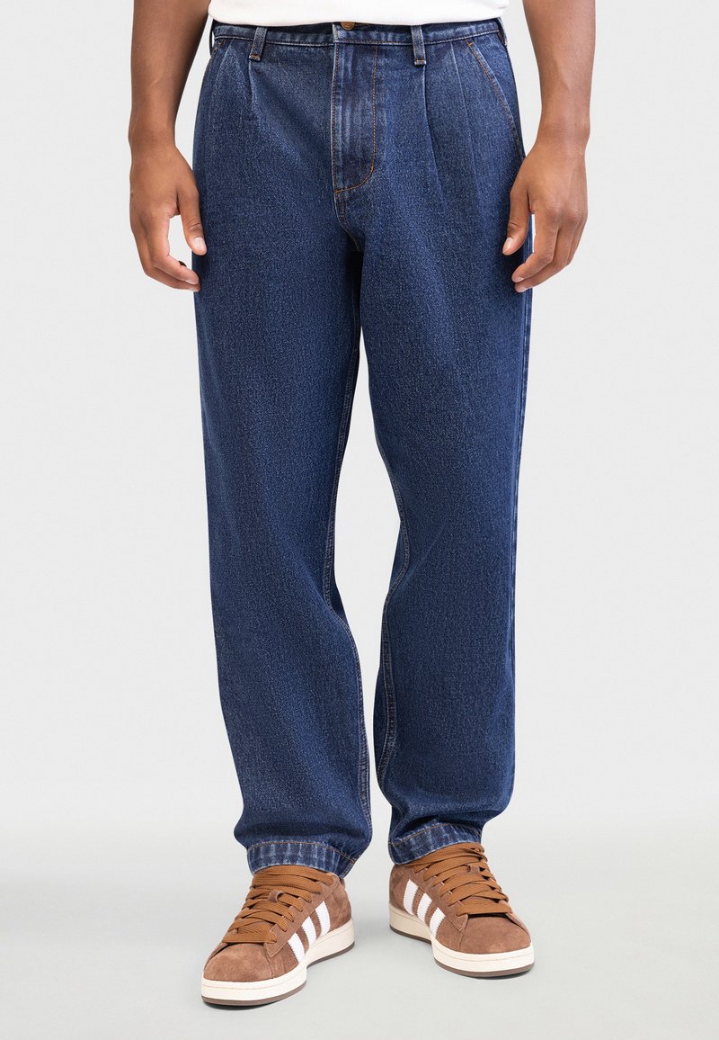 Tmavomodré rifle z denimu s rovnými nohavicami a prednými záhybmi. Kombinované s hnedými teniskami s bielymi pruhmi.