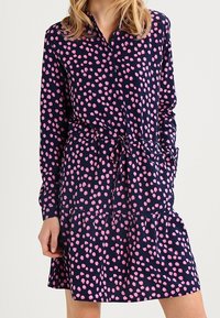 Marineblaues Kleid aus leichtem Stoff mit einem rosa gepunkteten Muster, langen Ärmeln, einer durchgehenden Knopfleiste und einem Kordelzug in der Taille.