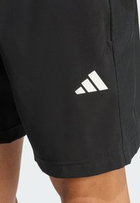 Pantalones cortos deportivos negros con un logo blanco de tres rayas de Adidas en el muslo izquierdo, llevados por una persona con piel bronceada visible.