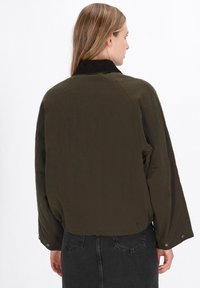Jachetă verde măslin cu guler din catifea, mâneci raglan și butoane prinse la manșete; textură netedă și croială lejeră.