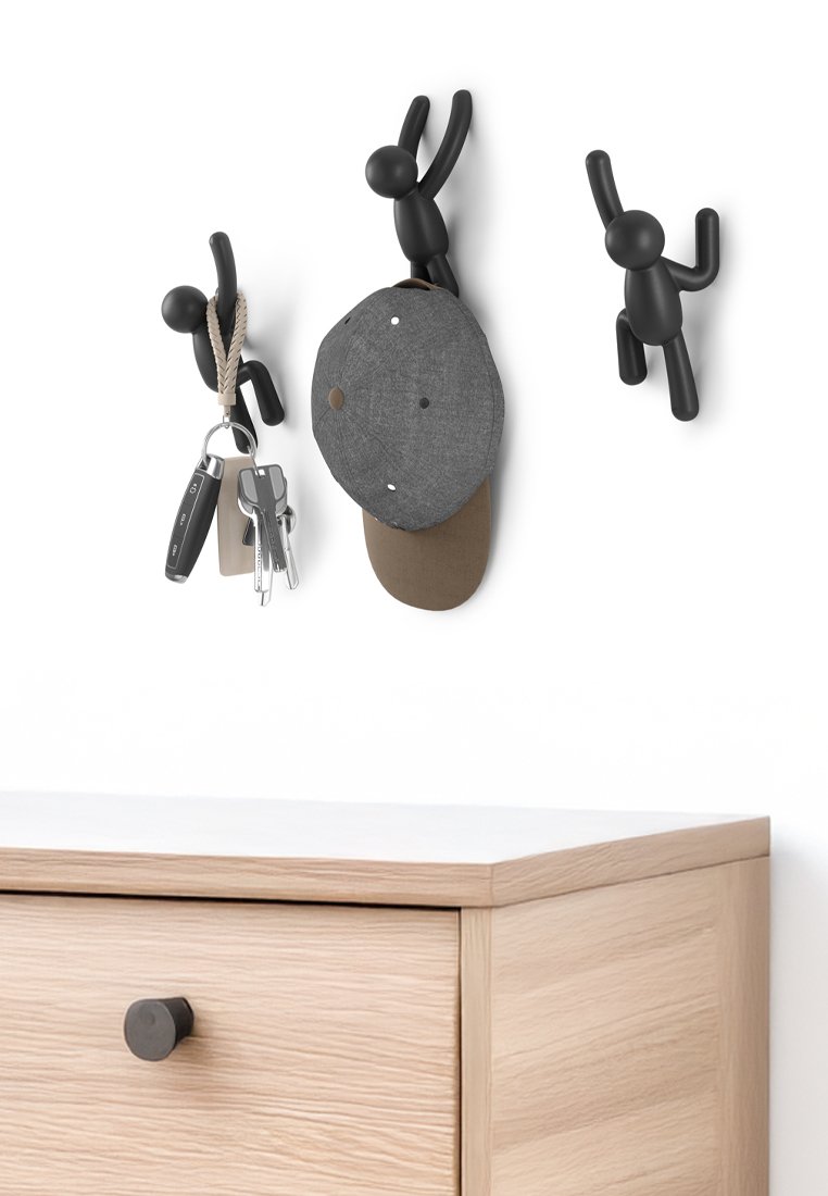 Ganci in plastica nera a forma di personaggi stilizzati montati su una parete, che tengono un cappello grigio e delle chiavi, sopra un mobiletto in legno chiaro.