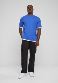 T-shirt bleu à manches courtes avec des accents blancs, associé à un pantalon cargo noir avec de multiples poches et des baskets blanches.