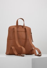 Sac à dos en cuir marron avec une finition texturée, de forme rectangulaire, deux bretelles réglables et une fermeture zippée en haut.