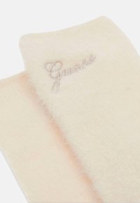 Heller, pelzähnlicher Schal mit weicher Textur, versehen mit silberner gestickter "Guess"-Logoschrift und Herzdetail. Glatte Kanten und leichter Glanz.