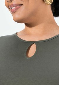 Olivgrön t-shirt med rundad halsringning och en nyckelhålsöppning. Tyget har en slät, stretchig textur.