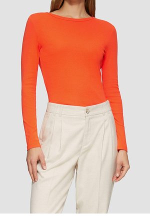 Vrouw draagt een nauwsluitend langarmig feloranje shirt dat in een high-waisted, geplooide lichtbeige broek is gestopt, tegen een effen achtergrond.