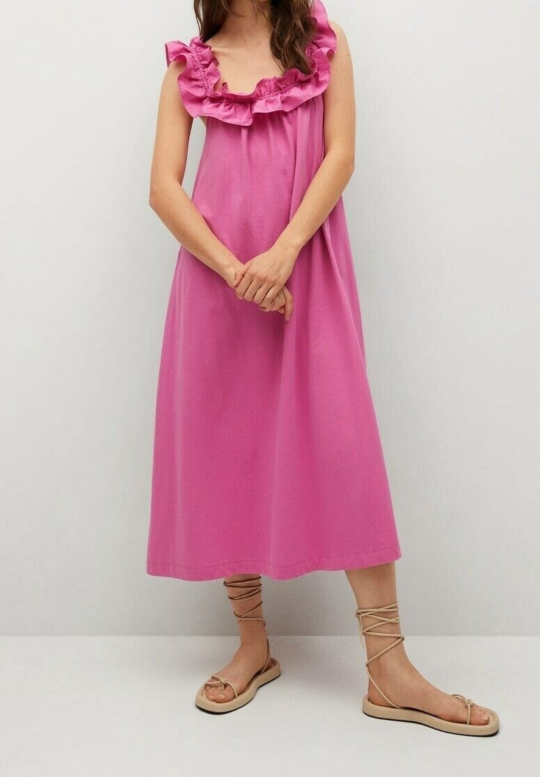 Femme debout, portant une robe midi sans manches rose vif avec un col à volants et des sandales plates beiges à lacets, sur un fond uni.