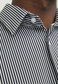 Camicia a righe bianche e nere con colletto abbottonato, caratterizzata da una trama liscia e sottili linee verticali, che evidenziano cuciture precise.