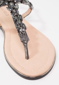 Musta koristeltu thong-sandaali, jossa on strassikoristeet, nahkaisella sisäpohjalla ja tyylikkäällä tasapohjalla. Kuosiset hihnat koristeellisilla yksityiskohdilla.