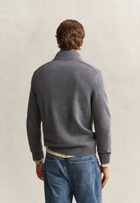 Mann trägt einen grauen gestrickten Pullover und blaue Jeans, von hinten vor einem einfachen hellen Hintergrund gezeigt.