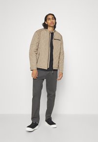 Beige vadderad jacka med hög krage, svart dragkedja och två sidofickor, buren över en svart topp, i kombination med grå byxor och svarta sneakers.