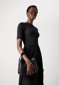 Donna che indossa un top nero attillato e una gonna plissettata, mentre tiene in mano una borsa a tracolla nera intrecciata, con un bracciale d'argento e orecchini a cerchio, su uno sfondo neutro.
