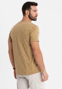Brun kortærmet t-shirt med et subtilt gentaget tekstmønster, rund halsudskæring, lavet af bomuldsstoffer og lige pasform. Etiketten er synlig på siden.