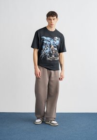 Juoda medvilninė marškinėliai su grafiniu dizainu, vaizduojančiu vilką ir žaibus. Derinama su laisvais šviesiai rudos spalvos sportiniais kelnėmis ir pilkais sportbačiais.