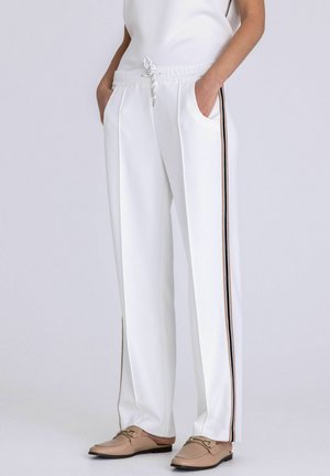 White relaxed-fit pants with front pleats and side black and beige stripes, worn with beige slip-on loafers and hands in pockets.