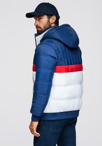 Padded jacket w niebiesko-białym wzorze z kolorowymi blokami, z czerwonymi akcentami. Ma kaptur i ściągacz na dole, o błyszczącej teksturze.