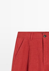 Rode broek met hoge taille, zijzakken, ceintuurlussen, plooien en een zwarte knoopsluiting in de taille.