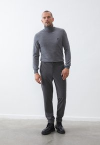 Pelēks turtlenecks džemperis ar logotipu, apvienots ar pelēkiem sašaurinātiem biksēm. Melni zābaki ar auklēm un izturīgiem zoleiem. Vienkāršs, mūsdienīgs dizains.