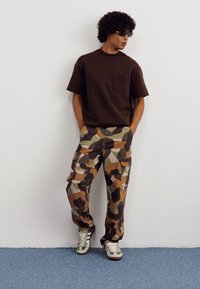 Ruskea lyhythihainen paita, jossa on rintatasku, yhdistettynä maastovärisiin cargo-housuihin. Kengät, joissa on vihreät ja valkoiset yksityiskohdat.