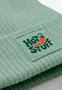 Light green knit beanie with a textured pattern. Features a square patch with the text "HOT STUFF" and a red chili pepper graphic.
