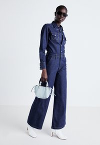 Salopetă din denim de culoare albastră închisă, cu închidere cu nasturi în față, buzunare la piept și picioare late. Accesoriile includ o geantă de umăr de culoare albastră deschisă și cizme cu toc bey.