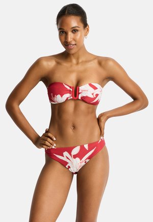 Donna che indossa un bikini rosso a fiori senza spalline con una chiusura a forma di U al centro, in piedi con una mano sul fianco su sfondo bianco.