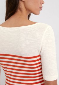 Crème gebreid top met korte mouwen, een wijd ronde halslijn en horizontale rood-oranje strepen over het onderste gedeelte. Soepele textuur.