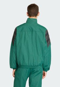 Grön lättviktsjacka med hög krage, svarta accenstrimlor på axlarna, elastiska muddvävar och en rynkad fåll för en avslappnad passform.