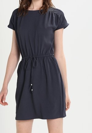 Frau trägt ein marineblaues, knielanges Kleid mit kurzen Ärmeln, einer taillierten Passform und einem Kordelzug, steht vor einem schlichten weißen Hintergrund.