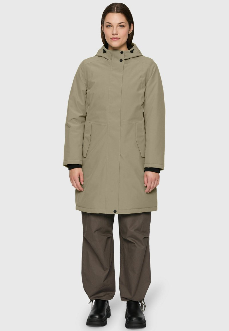Vrouw in een beige lange jas met capuchon, losse donkerbruine broek en zwarte laarzen, staande tegen een effen lichte achtergrond.