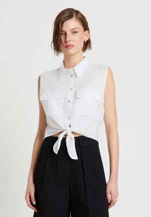 Woman with short hair wearing sleeveless white button-up shirt tied at waist and high-waisted black pants, standing against plain background.