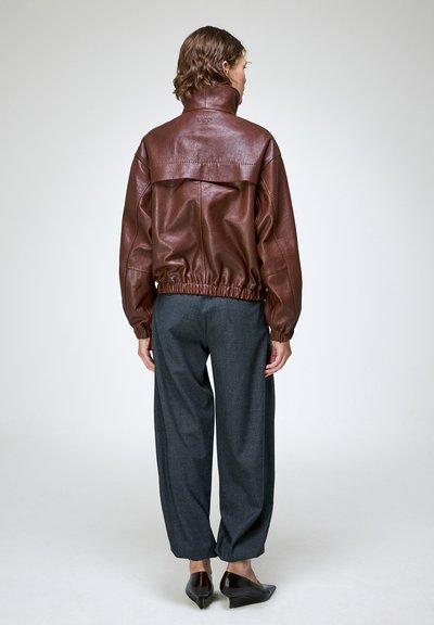Personne debout de dos, portant une veste en cuir marron foncé, un pantalon ample gris foncé et des chaussures à talons noires sur un fond uni.